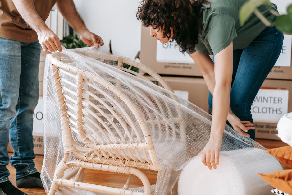 Two movers from Brompton Man and Van engaged in home relocation inside a house, carefully wrapping a rattan armchair with bubble wrap and securing it with plastic packaging material. One person, stand