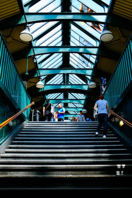 Inside a modern railway station, the image shows a wide staircase with metal handrails leading up to a glass-roofed walkway illuminated by natural light. Several individuals are ascending and descending the stairs; some are carrying backpacks or small bags, while others are walking empty-handed. The station's interior features yellow and blue structural elements on the ceiling and walls, with hanging spherical lights providing additional illumination. The environment appears busy, consistent with a typical urban station where people are commuting. This scene naturally relates to transportation and home relocation services, such as those offered by Brompton Man and Van, especially when coordinating the moving process through public transport or arranging furniture transport, packing, and loading activities.
