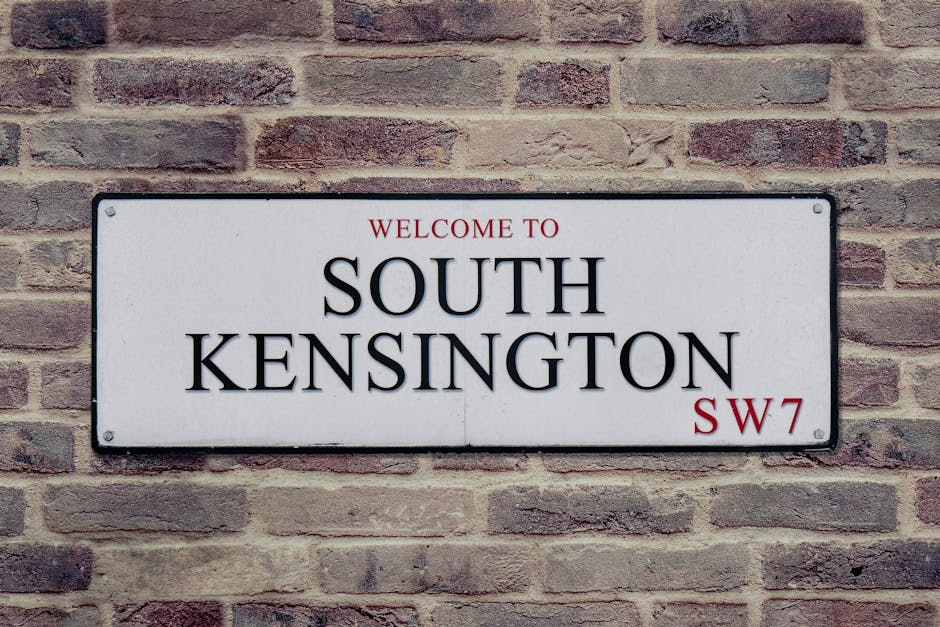 A rectangular metal street sign mounted on a brick wall displaying the words 'Welcome to South Kensington' in black capital letters, with 'South Kensington' larger and central. The sign has a white background with black border and red text indicating 'SW7' in the bottom right corner. The brick wall behind the sign is composed of reddish-brown bricks with visible mortar joints and some variation in brick shade. The lighting appears natural, highlighting the texture of both the bricks and the sign. This image relates to home relocation or moving services, illustrating an entry point or notable location as part of a furniture transport or packing and moving process managed by Brompton Man and Van, a company specialising in removals in the South Kensington area, supporting efficient and organised house removals.