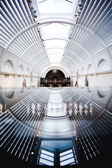 A spacious interior view of the Victoria and Albert Museum featuring a high, curved glass ceiling with bright natural light streaming through. The ceiling is supported by white, evenly spaced steel ribs, creating a symmetrical pattern above. The image captures the long corridor with white walls and decorative sculptures placed along the sides. The polished, reflective floor creates a mirror-like effect, showing an inverted reflection of the ceiling structure and the corridor below. The environment appears clean and well-lit, emphasizing the architectural detail of the museum's interior, which is relevant to house removals and moving services focused on precise packing and careful transport of valuable artworks or furniture within cultural institutions or private homes heading for collections or exhibitions. Brompton Man and Van occasionally associates such interior spaces with professional removal and furniture transport processes involved in home relocation or institutional moves.